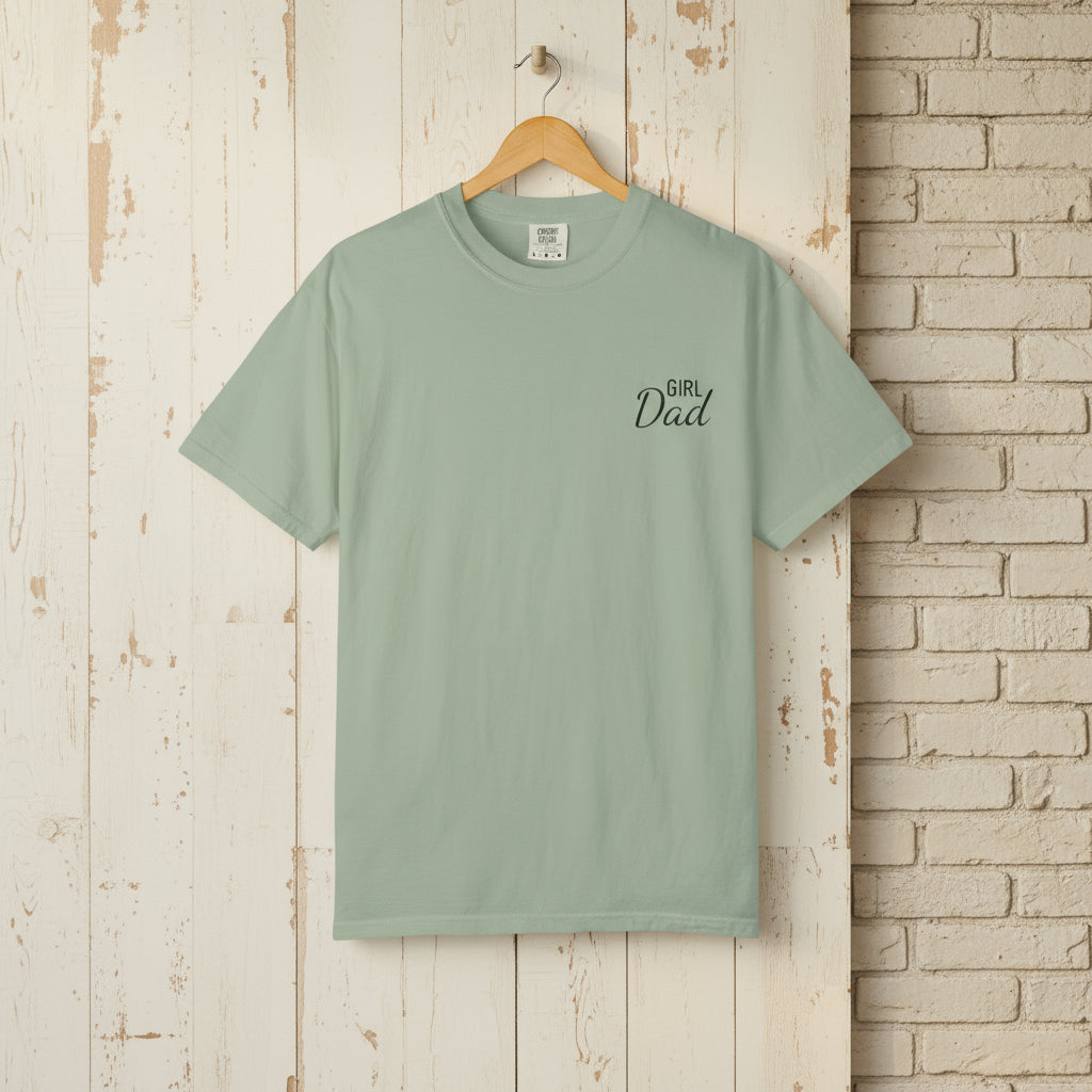Mint green t-shirt with 'Girl Dad' text on a wooden hanger against a rustic brick wall.