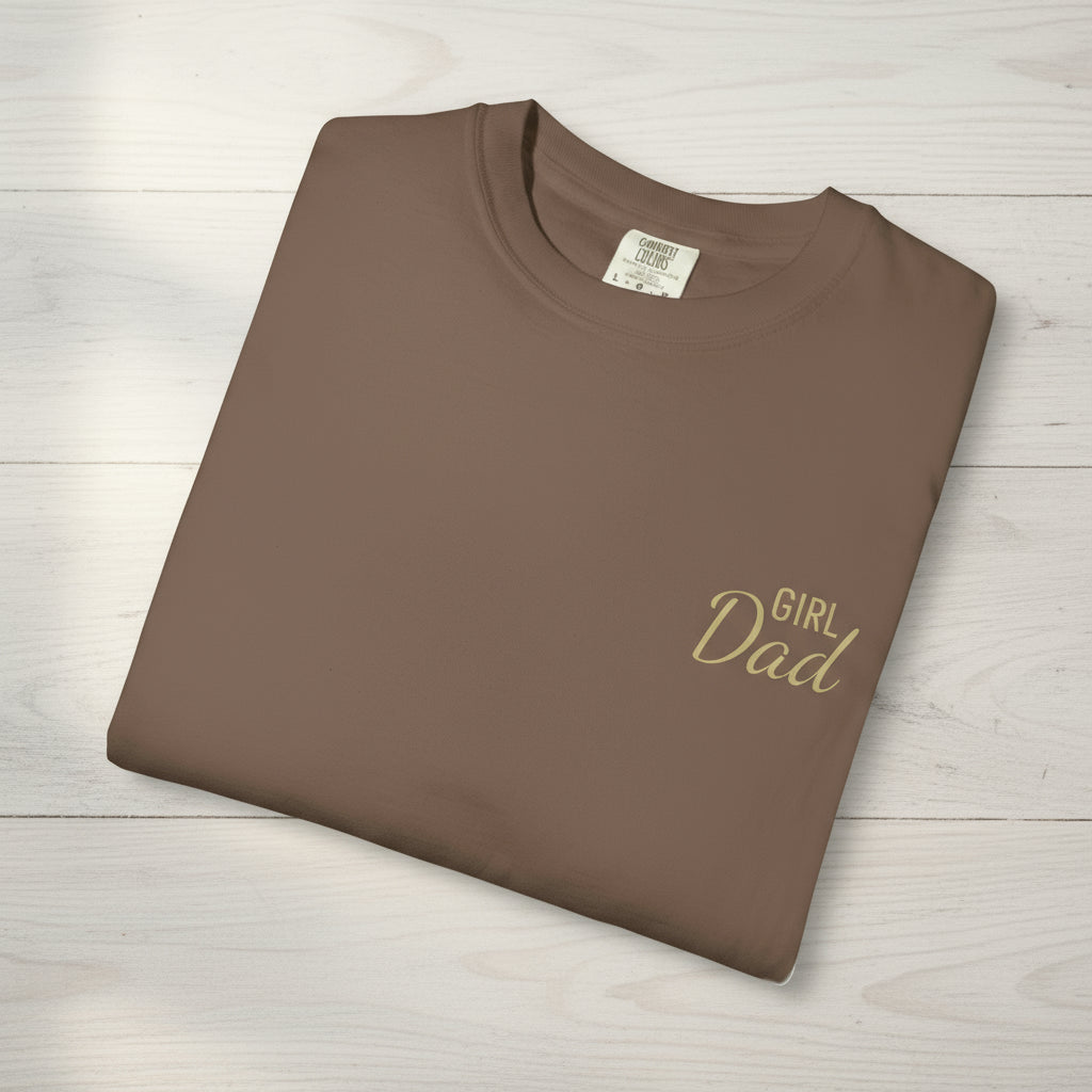 Folded brown t-shirt with 'Girl Dad' text on a light wooden surface