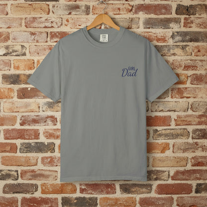 Gray t-shirt with 'Girl Dad' text on a wooden hanger against a brick wall.