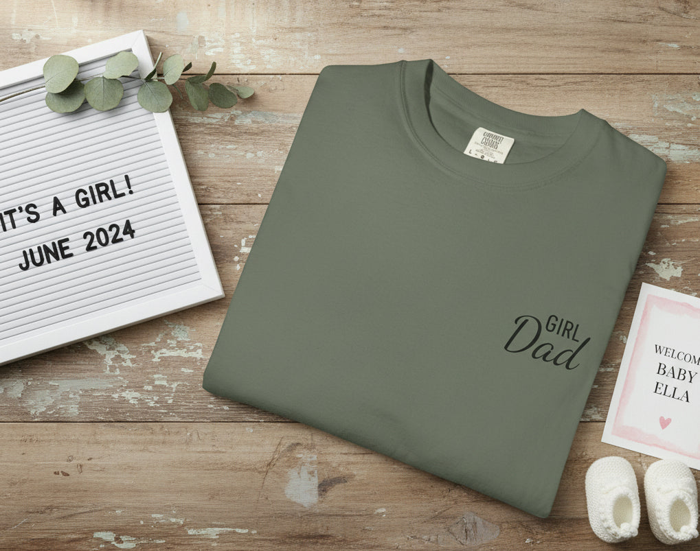 Green t-shirt with 'Girl Dad' text on a wooden surface with pregnancy announcement items.