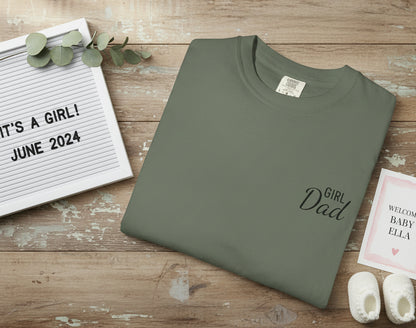 Green t-shirt with 'Girl Dad' text on a wooden surface with pregnancy announcement items.